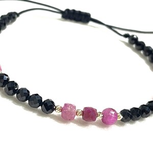 Op de afbeelding: Verstelbare armband met afwisselend zwarte facetkralen en roze edelsteen kralen. De armband heeft een zwart koord en goudkleurige accenten. De roze kralen zijn een mix van ronde en facetvormen.