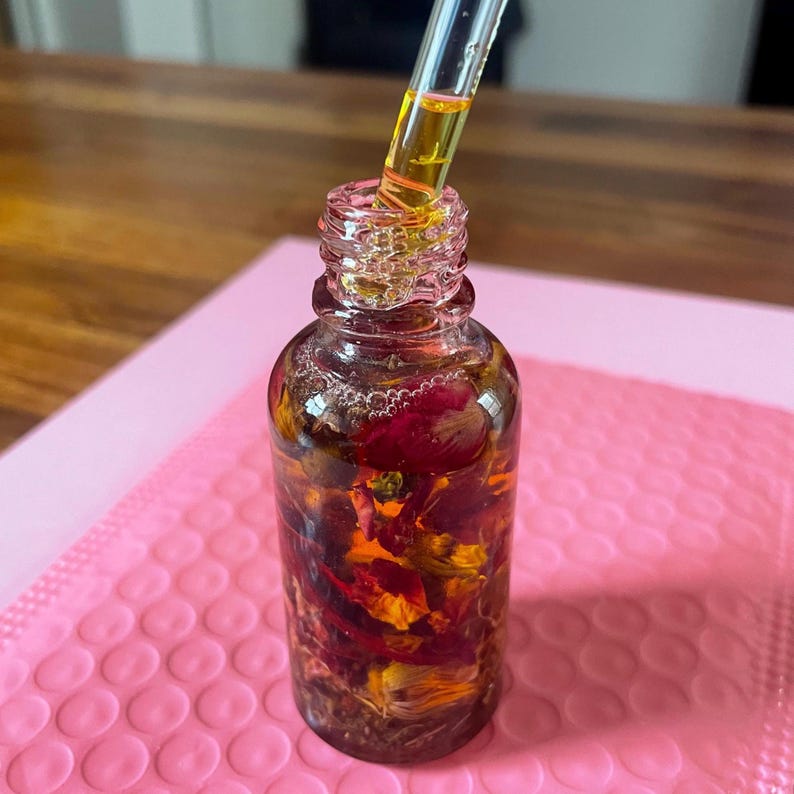 May include: A clear glass bottle containing a golden liquid and dried flowers, with a glass dropper. The bottle is on a pink surface, with a wooden surface behind it.