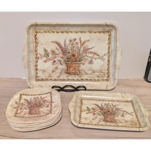 May include: A set of serving trays and square plates featuring a cream-colored background with a marble-like texture and a central illustration of a floral arrangement in a woven basket, framed by a decorative border with a rope-like pattern.