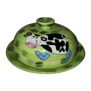 May include: A round ceramic butter dish with a dome-shaped lid featuring a light green background with dark green brushstrokes. A cartoon cow with black and white spots, wearing blue boots, is painted on the center of the lid.