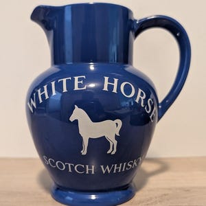 May include: A glossy dark blue ceramic pitcher featuring a white silhouette of a horse in the center. The text "WHITE HORSE" is printed in an arc above the horse, and the words "SCOTCH WHISKY" are printed in a straight line below the horse.