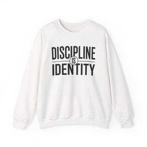 Peut inclure: Un sweat-shirt blanc à manches longues et col rond avec le texte noir DISCIPLINE IS IDENTITY imprimé sur la poitrine. Le mot DISCIPLINE est sur la ligne supérieure, le mot IS est dans un cercle au milieu et IDENTITY est en bas.