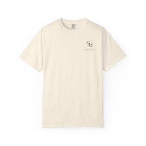 Peut inclure: Un t-shirt à manches courtes de couleur crème avec une encolure ronde. Sur la poitrine supérieure gauche, un logo noir présente les lettres SM empilées verticalement, avec le texte SILENT MOVES imprimé en dessous dans une police sans empattement.