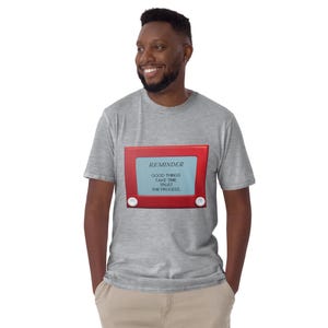 May include: A light gray t-shirt featuring a red framed graphic that resembles an Etch-A-Sketch. The graphic has the word "REMINDER" at the top, with the words "GOOD THINGS TAKE TIME TRUST THE PROCESS" below. The shirt is being worn by a person.