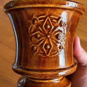 May include: A brown ceramic vase with an embossed floral design. The vase features a flared top, a rounded base, and a pedestal foot. The exterior has a glossy finish.