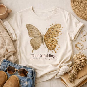 Pode incluir: Camiseta creme com uma ilustração detalhada de uma borboleta, com uma asa em tom natural e a outra dourada. O texto "The Unfolding" e uma citação estão impressos abaixo. A camiseta está dobrada.