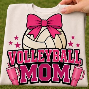 Könnte beinhalten: Ein cremefarbenes T-Shirt mit einer Grafik eines Volleyballs, der von einer großen rosa Schleife gekrönt wird. Unter dem Ball ist der Text VOLLEYBALL MOM in fetten rosa Buchstaben gedruckt, flankiert von zwei rosa Bechern und kleinen Sternen.