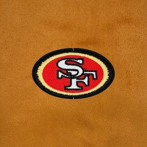 Puede incluir: Una superficie de tela de color dorado con un parche bordado de forma ovalada. El parche muestra las letras SF en blanco con un contorno negro sobre un fondo rojo, todo ello encerrado en un borde amarillo y un borde exterior negro grueso.