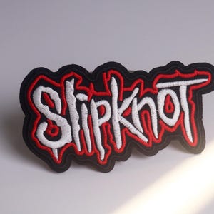 Puede incluir: Parche bordado con el nombre de la banda "Slipknot" en blanco, contorneado en rojo, sobre un fondo negro. El parche tiene un borde dentado y es para ropa o accesorios.