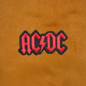 Pode incluir: Uma superfície de tecido amarelo mostarda com um emblema bordado ao centro. O emblema exibe o logótipo vermelho AC/DC com um raio central, contornado com fio preto. O bordado tem uma textura em relevo sobre o tecido liso de fundo.