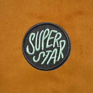Puede incluir: Un parche bordado circular que presenta la palabra SUPERSTAR en hilo verde menta claro sobre un fondo negro. El parche está centrado sobre una superficie de tela de color amarillo mostaza, texturizada y de aspecto mate.