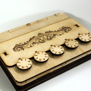 May include: A wooden box with a decorative floral design and five circular dials with letters. The box is made of light-colored wood and has a hinged lid. The dials are arranged in a row and appear to be part of a locking mechanism.