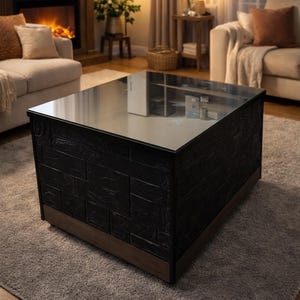 Puede incluir: Una mesa de centro cuadrada moderna con un exterior negro texturizado y una tapa de cristal. La base es de color madera oscura. La mesa está sobre una alfombra gris en un salón.