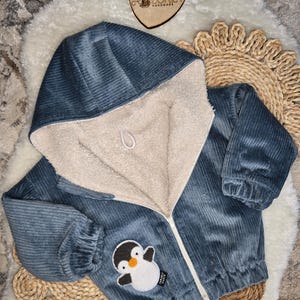 Könnte beinhalten: Blaue Babyjacke aus Cord mit Kapuze und cremefarbenem Fleecefutter. Die Jacke hat einen Reißverschluss und einen Pinguin-Aufnäher. Sie liegt auf einer weißen, flauschigen Oberfläche.