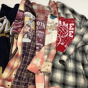 May include: A collection of six long-sleeved shirts arranged side-by-side. The garments include a navy blue shirt with yellow plaid accents, a purple plaid shirt with a rose graphic, a plaid shirt with a cowgirl print, a pink plaid shirt with text, a light pink plaid shirt with a butterfly graphic, and a black and white plaid shirt with a red graphic featuring a volleyball and the text GAME DAY.