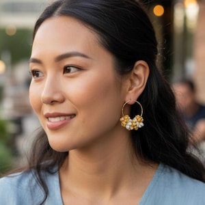 May include: Gold hoop earrings with clusters of yellow and white beads. The earrings are worn by a person with dark hair and a blue top. The background is blurred, suggesting an outdoor setting.