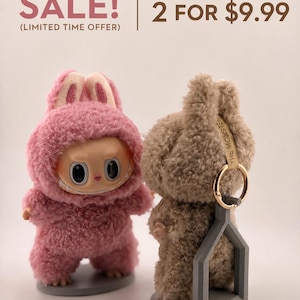May include: Two plush monster figures, one pink and one tan, each wearing a fuzzy hooded suit with rabbit-like ears. The tan figure has a gold keyring attached to its head and is supported by a gray plastic stand. Text reads: SALE! (LIMITED TIME OFFER) 2 FOR $9.99.