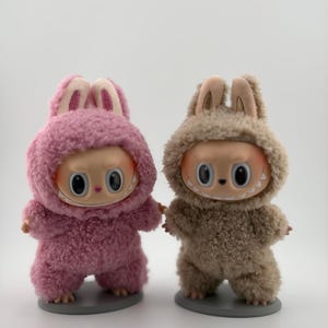 May include: Two small figurines standing on circular gray bases. One figurine wears a fuzzy pink hooded suit with rabbit ears, and the other wears a fuzzy beige hooded suit with rabbit ears. Both have large eyes and wide, toothy grins.