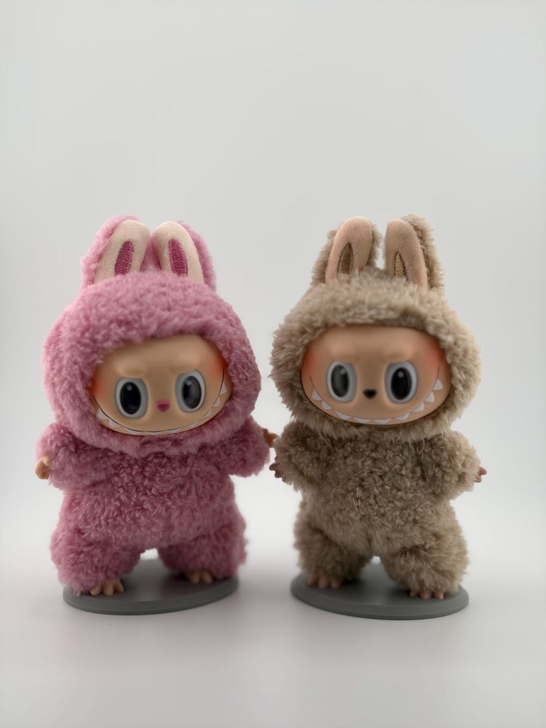 May include: Two small figurines standing on circular gray bases. One figurine wears a fuzzy pink hooded suit with rabbit ears, and the other wears a fuzzy beige hooded suit with rabbit ears. Both have large eyes and wide, toothy grins.