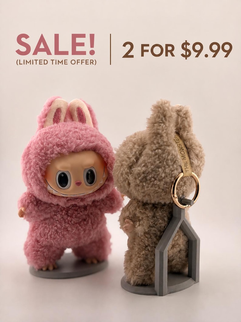May include: Two plush monster figures, one pink and one tan, each wearing a fuzzy hooded suit with rabbit-like ears. The tan figure has a gold keyring attached to its head and is supported by a gray plastic stand. Text reads: SALE! (LIMITED TIME OFFER) 2 FOR $9.99.