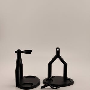 May include: Two black plastic display stands with circular bases and vertical supports featuring a top ring for holding items, accompanied by a separate small black plastic Y-shaped component resting on the surface in front of the stands.