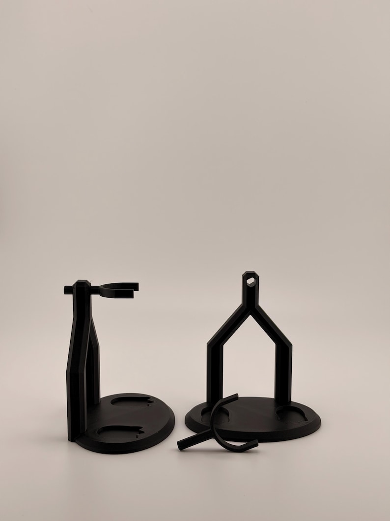May include: Two black plastic display stands with circular bases and vertical supports featuring a top ring for holding items, accompanied by a separate small black plastic Y-shaped component resting on the surface in front of the stands.