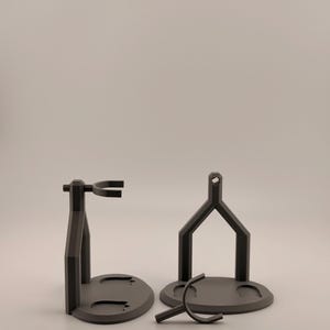 May include: Two matte dark gray display stands with circular bases and vertical support structures. One stand is fully assembled with a horizontal cradle, while the other is partially disassembled with a separate U-shaped component in front.