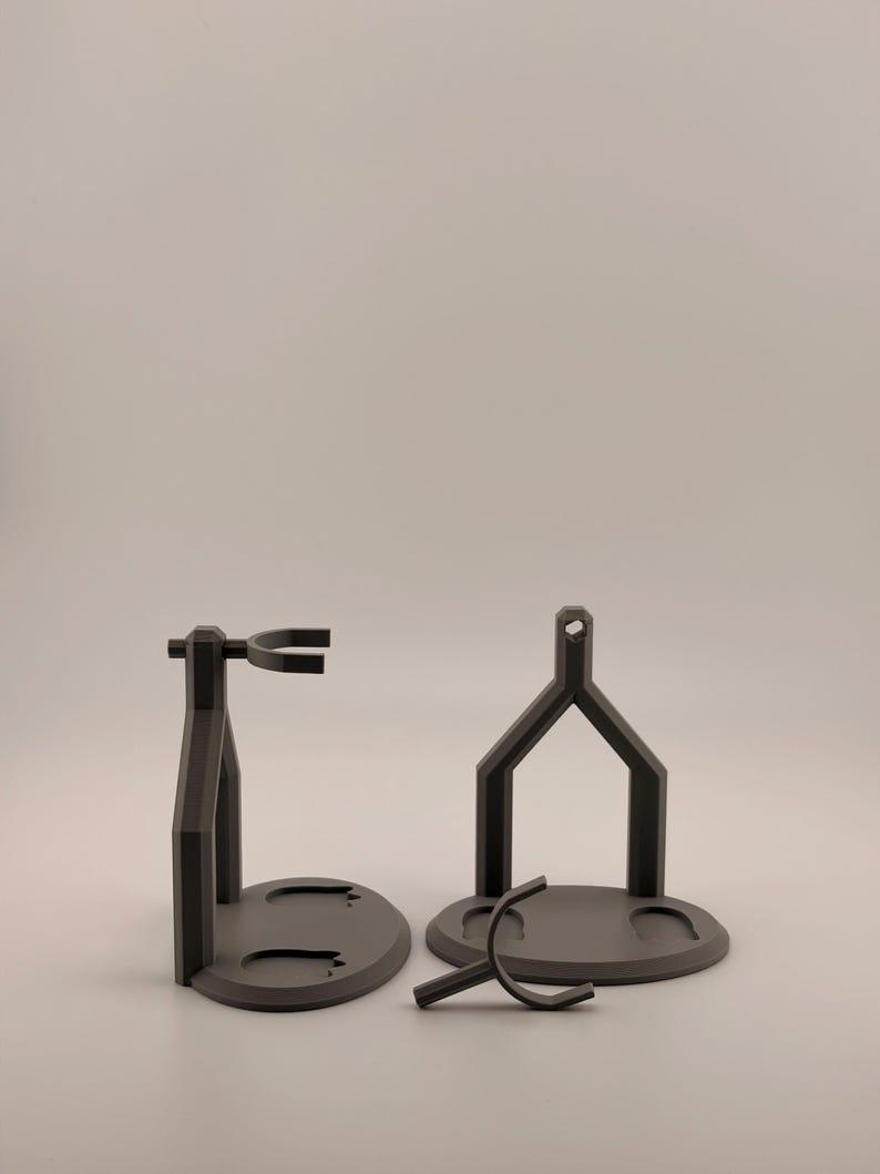 May include: Two matte dark gray display stands with circular bases and vertical support structures. One stand is fully assembled with a horizontal cradle, while the other is partially disassembled with a separate U-shaped component in front.