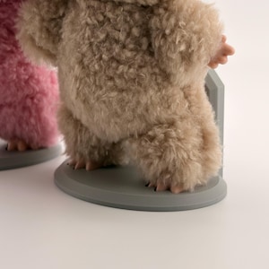 May include: A beige fuzzy creature figurine with a visible mouth and white teeth, standing on a round gray base. The figure is covered in a textured, shaggy material and is positioned next to a partial view of a pink fuzzy creature figurine.