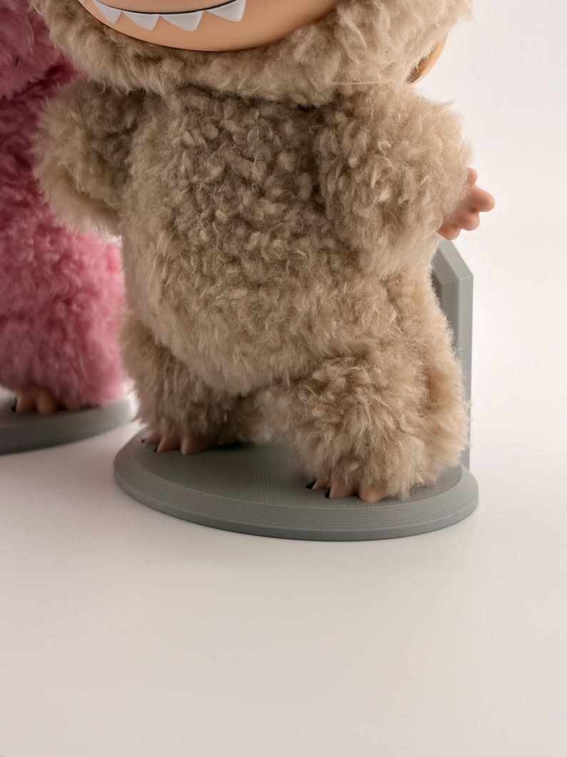 May include: A beige fuzzy creature figurine with a visible mouth and white teeth, standing on a round gray base. The figure is covered in a textured, shaggy material and is positioned next to a partial view of a pink fuzzy creature figurine.