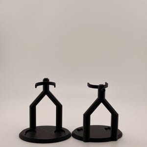 May include: Two black display stands with circular bases and Y-shaped supports. Each stand features a top cradle designed to hold an object securely. The structures are matte black and have a uniform, geometric design with a stable, flat base.