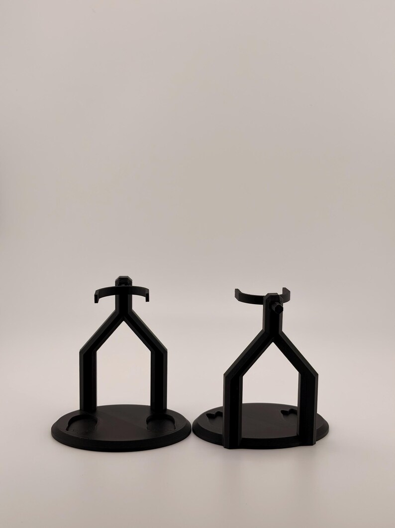 May include: Two black display stands with circular bases and Y-shaped supports. Each stand features a top cradle designed to hold an object securely. The structures are matte black and have a uniform, geometric design with a stable, flat base.
