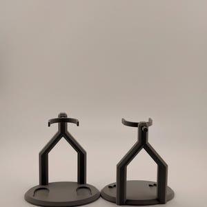 May include: Two matte black stands designed for holding objects. Each stand features a circular base and a Y-shaped support structure that extends upward to a curved cradle. The left stand has two small circular indentations on its base.