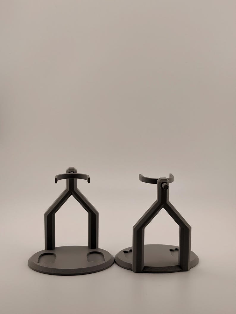 May include: Two matte black stands designed for holding objects. Each stand features a circular base and a Y-shaped support structure that extends upward to a curved cradle. The left stand has two small circular indentations on its base.