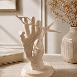 Puede incluir: Una escultura decorativa de mano de color crema que sostiene unas tijeras plateadas. La escultura está sobre una base redonda. Las tijeras están abiertas. El fondo incluye un jarrón con flores secas.