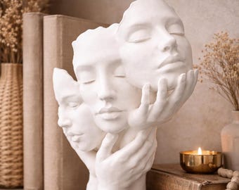 Peut inclure: Une sculpture blanche mate représentant trois visages humains entrelacés avec des mains sculptées. Le visage central est flanqué de deux visages plus petits, chacun soutenu par des mains détaillées. L'objet est fait d'un matériau lisse et solide.
