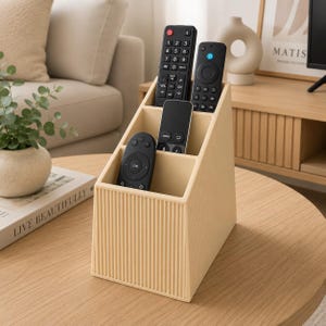 Puede incluir: Un organizador de mandos a distancia de plástico beige con un diseño escalonado y una textura exterior de estrías verticales. El soporte cuenta con cuatro compartimentos distintos que contienen cuatro mandos a distancia electrónicos negros de varios tamaños.