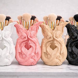 May include: Four mermaid-shaped makeup brush holders in white, pink, beige, and black. Each holder is filled with makeup brushes. The holders are on a white surface.