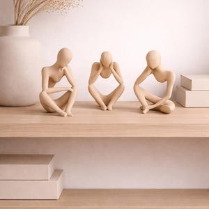 Pode incluir: Três esculturas minimalistas de figuras sentadas em tons neutros. Cada figura está posada de forma diferente, com as mãos sustentando a cabeça ou o queixo. As esculturas são exibidas em uma prateleira de madeira clara, com um vaso e uma planta seca ao fundo.