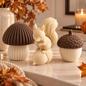 May include: A set of three decorative figurines featuring a brown mushroom with a ribbed cap, a cream-coloured squirrel perched on an acorn, and a cream-coloured acorn with a textured brown cap. Each piece is crafted with detailed surface textures.
