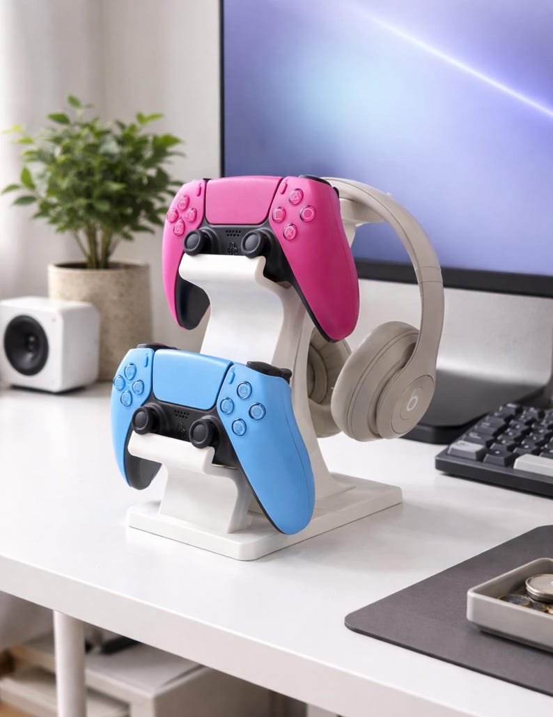 May include: A white gaming controller stand holding a pink and a blue controller, with a pair of gray headphones. The controllers are designed with black accents and buttons. The stand is on a white desk.
