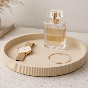 May include: A neutral-toned decorative tray holds a gold wristwatch, a clear perfume bottle with a gold cap, and a delicate gold bracelet. A small vase with dried flowers and a book are in the background.