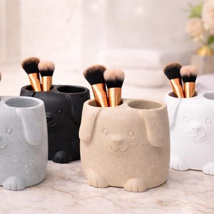 Puede incluir: Varios soportes de cerámica con forma de perro en gris, negro, beige y blanco, cada uno con dos compartimentos. Algunos contienen brochas de maquillaje, otros un cepillo de dientes. Diseño de perro de dibujos animados.