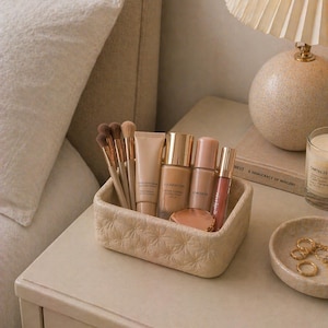 K&ouml;nnte beinhalten: Ein beigefarbener, strukturierter rechteckiger Beh&auml;lter enth&auml;lt Make-up-Pinsel, Foundation, Concealer und Lipgloss. Der Beh&auml;lter steht auf einem cremefarbenen Nachttisch neben einer kleinen Schale mit goldenen Ringen und einer brennenden Kerze.