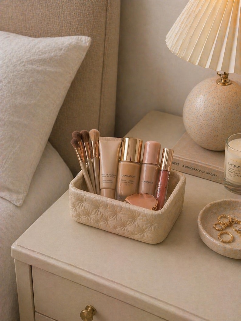 K&ouml;nnte beinhalten: Ein beigefarbener, strukturierter rechteckiger Beh&auml;lter enth&auml;lt Make-up-Pinsel, Foundation, Concealer und Lipgloss. Der Beh&auml;lter steht auf einem cremefarbenen Nachttisch neben einer kleinen Schale mit goldenen Ringen und einer brennenden Kerze.