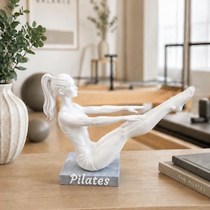 Peut inclure: Statuette blanche de Pilates en position de yoga, mont&eacute;e sur une base grise avec le mot "Pilates" en blanc. La figurine est sur une surface en bois, avec un vase et une image encadr&eacute;e en arri&egrave;re-plan.