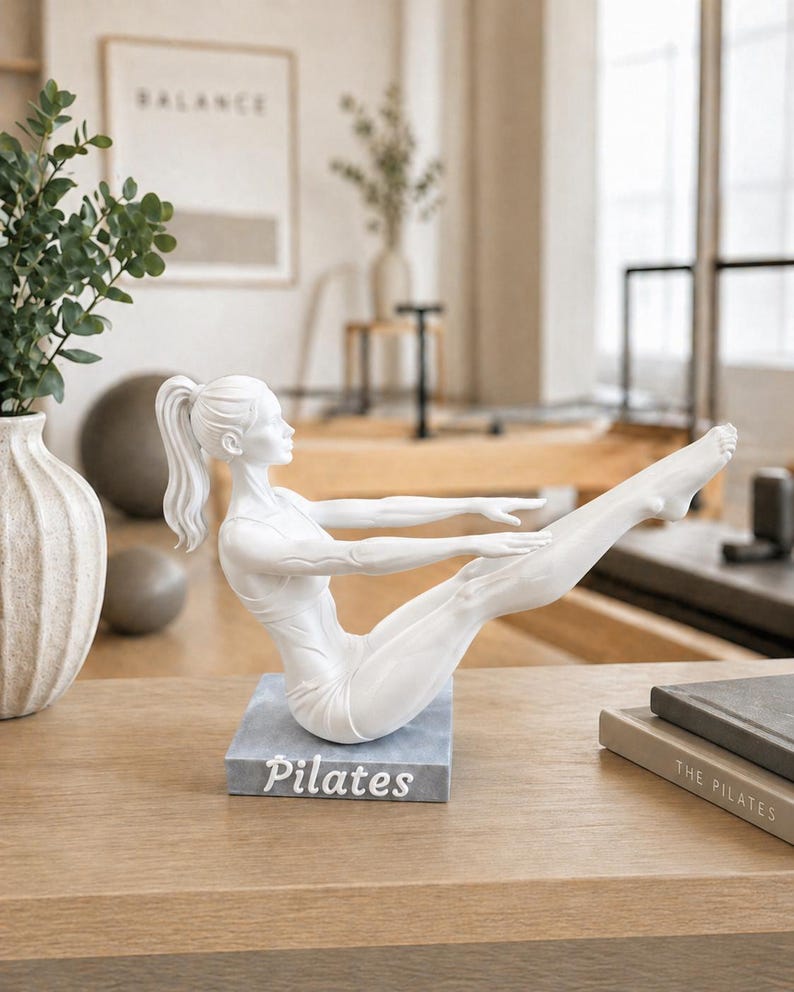 Peut inclure: Statuette blanche de Pilates en position de yoga, mont&eacute;e sur une base grise avec le mot "Pilates" en blanc. La figurine est sur une surface en bois, avec un vase et une image encadr&eacute;e en arri&egrave;re-plan.