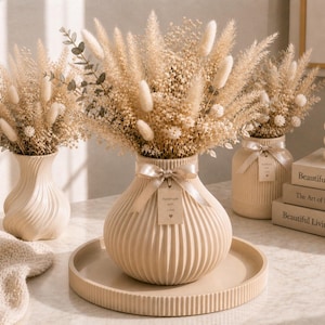 Op de afbeelding: Drie beige keramische vazen met droge bloemstukken. De vazen staan op een beige dienblad en een tafel met boeken en een kaars. De middelste vaas heeft een lint en een label met de tekst "Handmade with love".