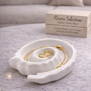 May include: A white, shell-shaped decorative tray holds gold jewellery, including a necklace and rings. The tray sits on a marble surface, with a box labelled "Rovera Selections" in the background. The theme is modern home decor.