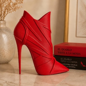 May include: A vibrant red high-heeled boot with a pointed toe and stiletto heel. The boot features a unique design with angular lines and a high collar. The heel is approximately 5 inches tall. The book titles "EL DIABLO VISTE A LA MODA" are visible in the background.